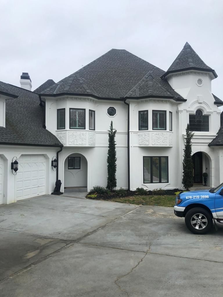 A two-story white house with dark shingled roofs and a turret, with a parked blue vehicle in the foreground.