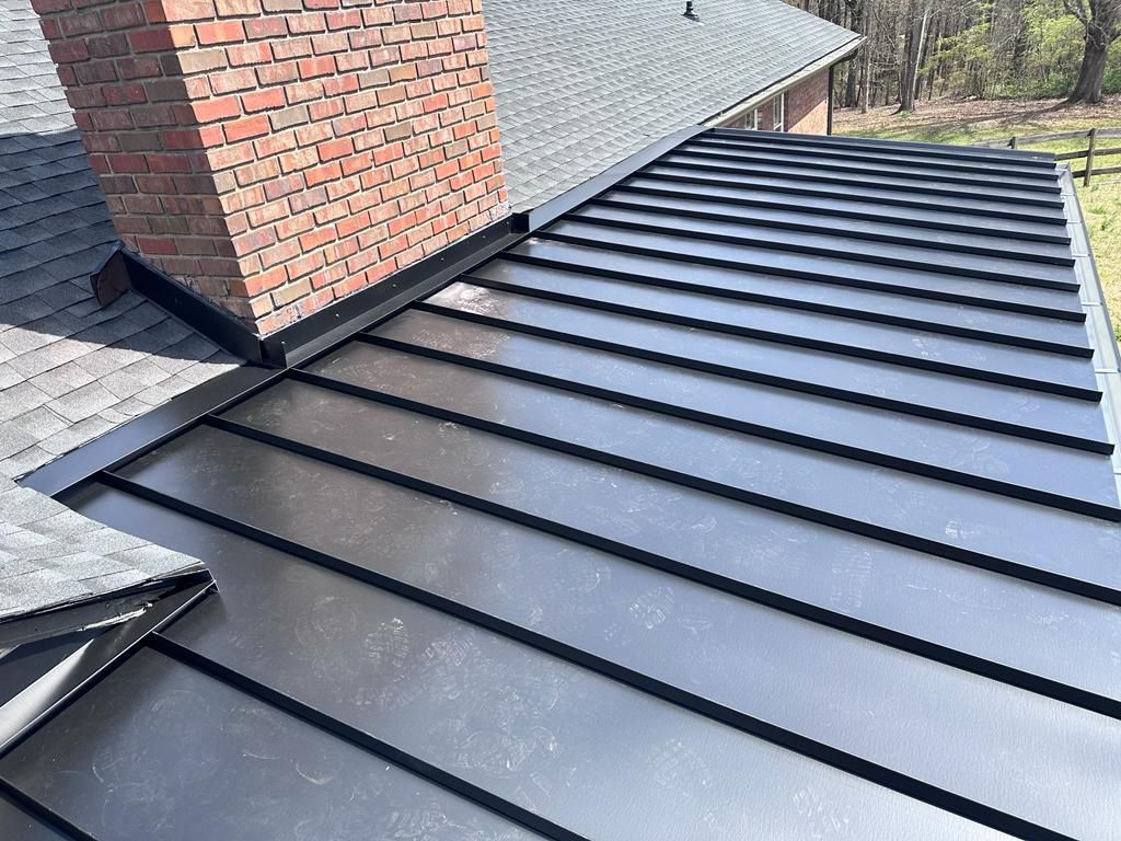 A metal roof with vertical standing seams meets a brick chimney and an asphalt shingle roof section.