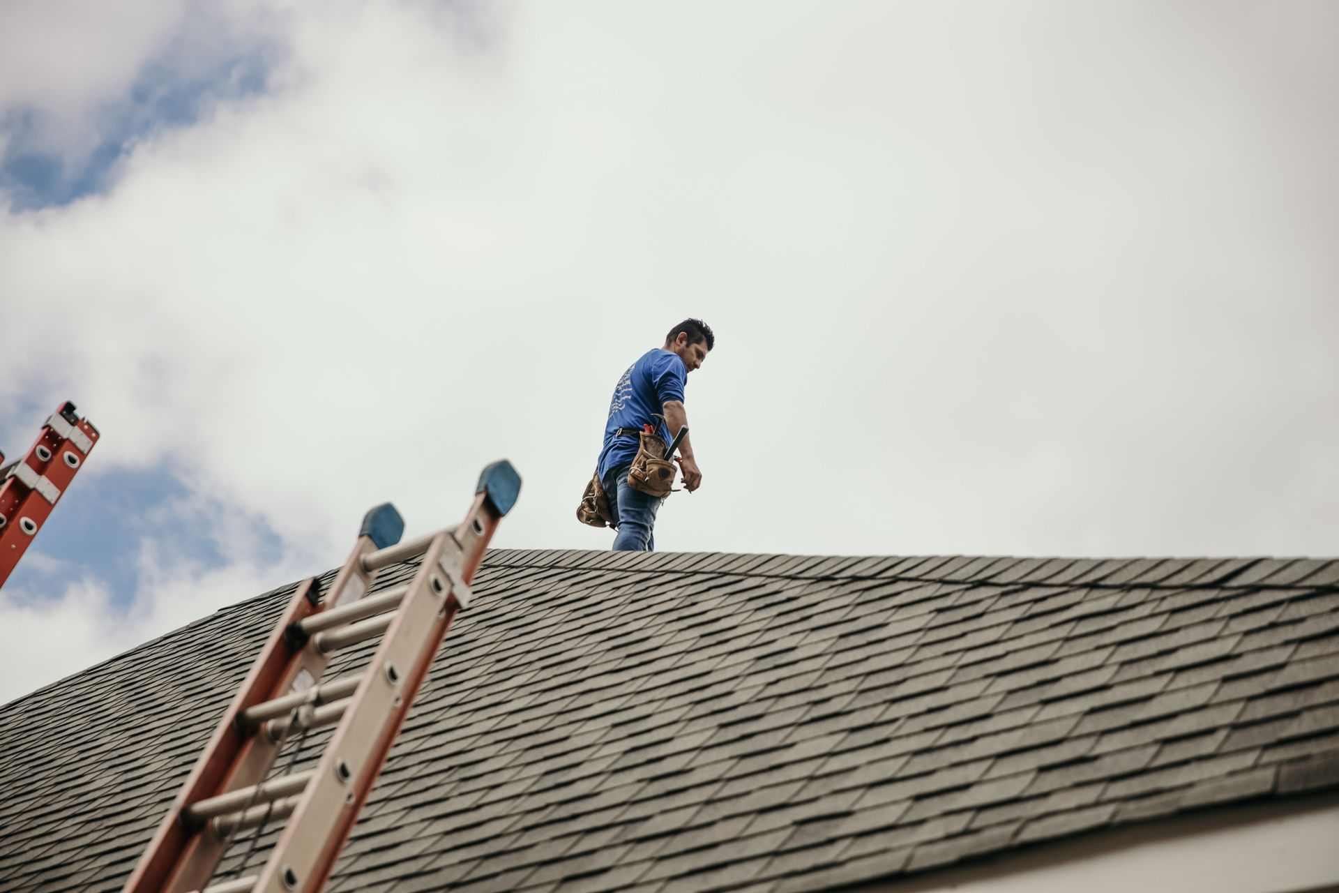 A worker in a blue shirt and tool belt stands on a shingled roof next to an extended ladder against a cloudy sky.