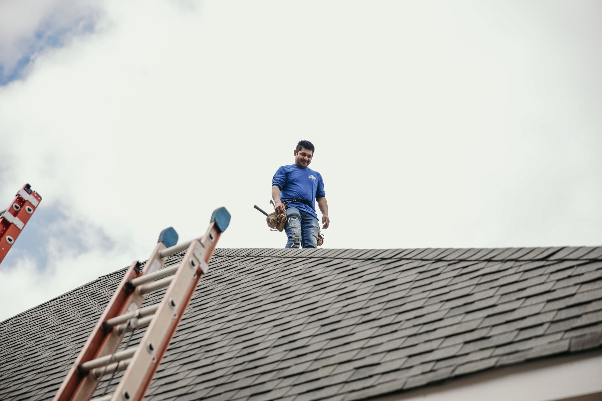 A person in a blue shirt and tool belt stands on a shingled roof next to an extended ladder.