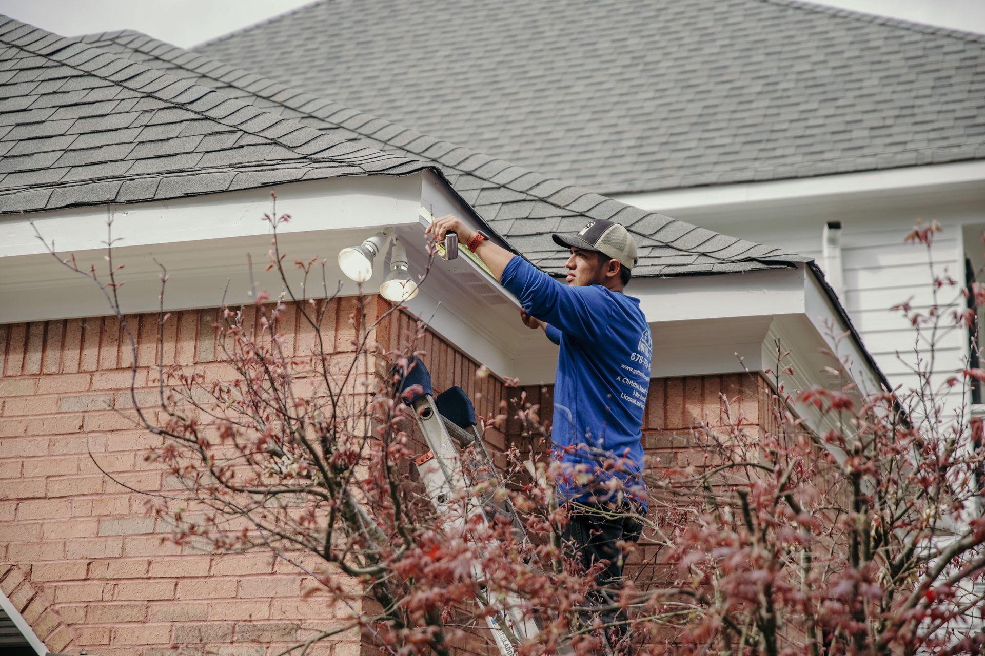 Two workers in blue shirts walk across a shingled residential roof under a cloudy sky.