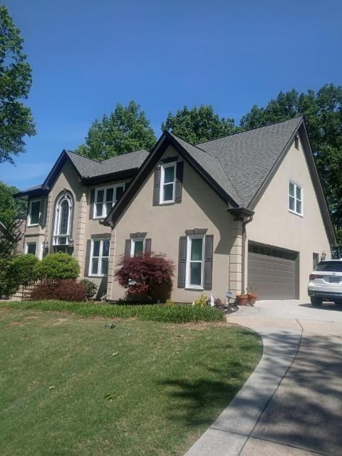 A two-story, tan-colored house with a brown roof and shutters, a front lawn, and a driveway with a parked white SUV.