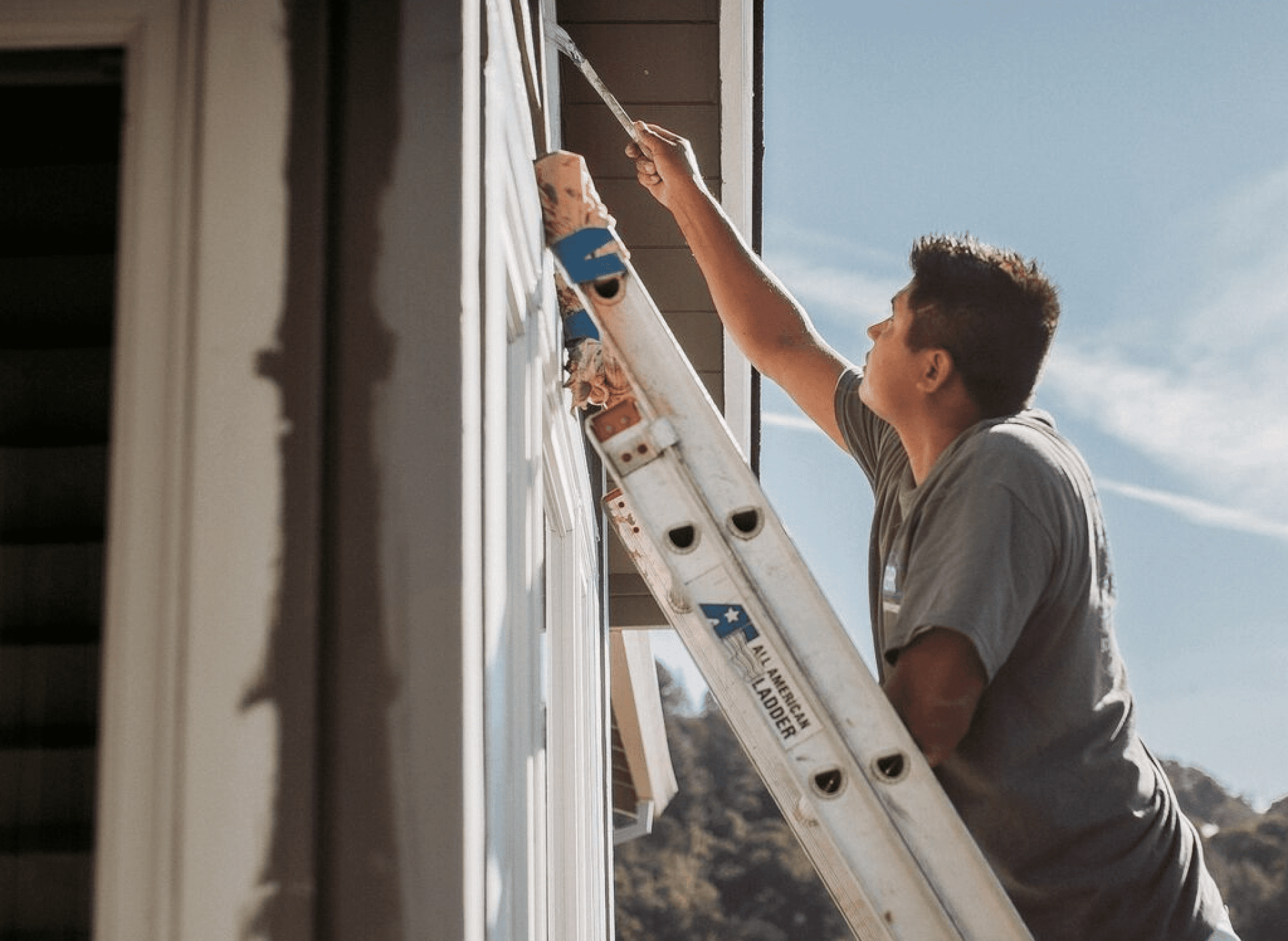 A person standing on an extension ladder paints the exterior trim of a house.