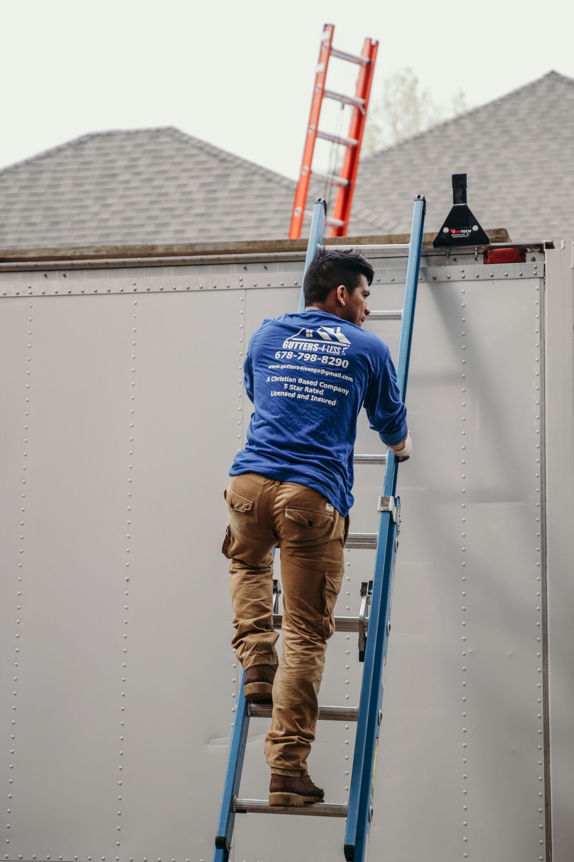 A person in a blue long-sleeved shirt and tan pants climbing a blue ladder against a white trailer.