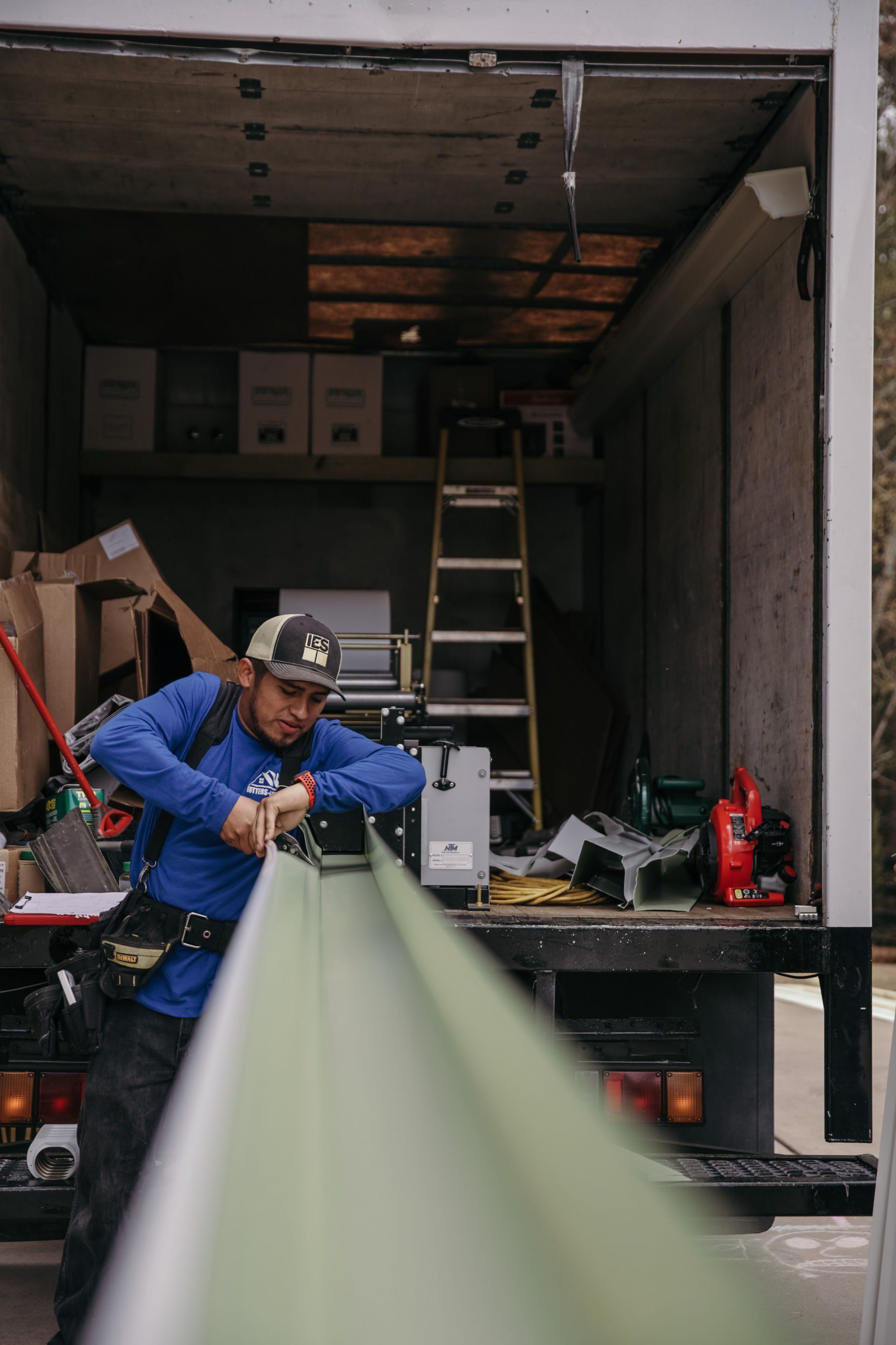 A worker in a blue long-sleeve shirt operates a gutter machine inside the back of a box truck.