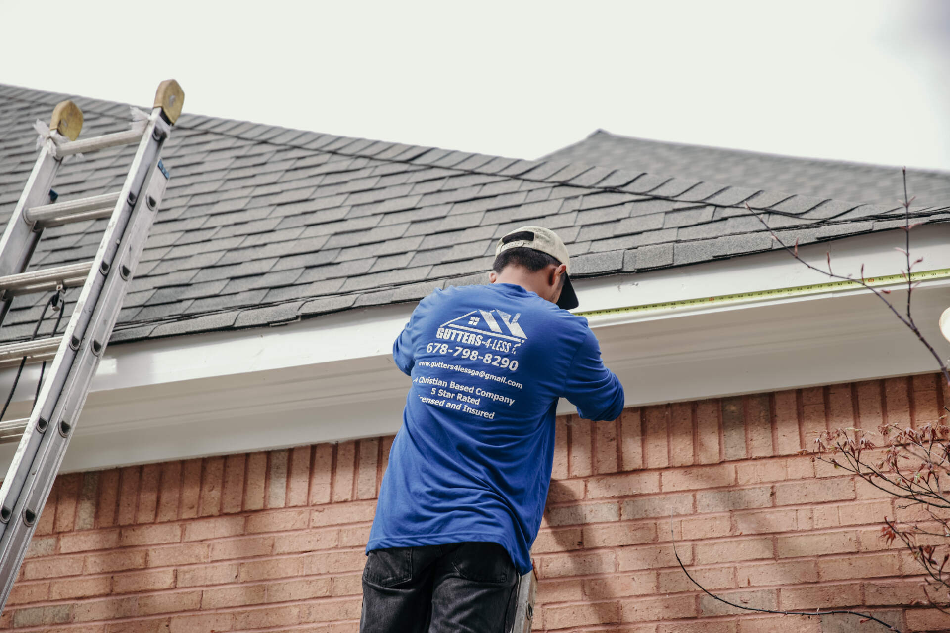 A person in a blue long-sleeved shirt works on the white rain gutters of a brick house near a metal ladder.