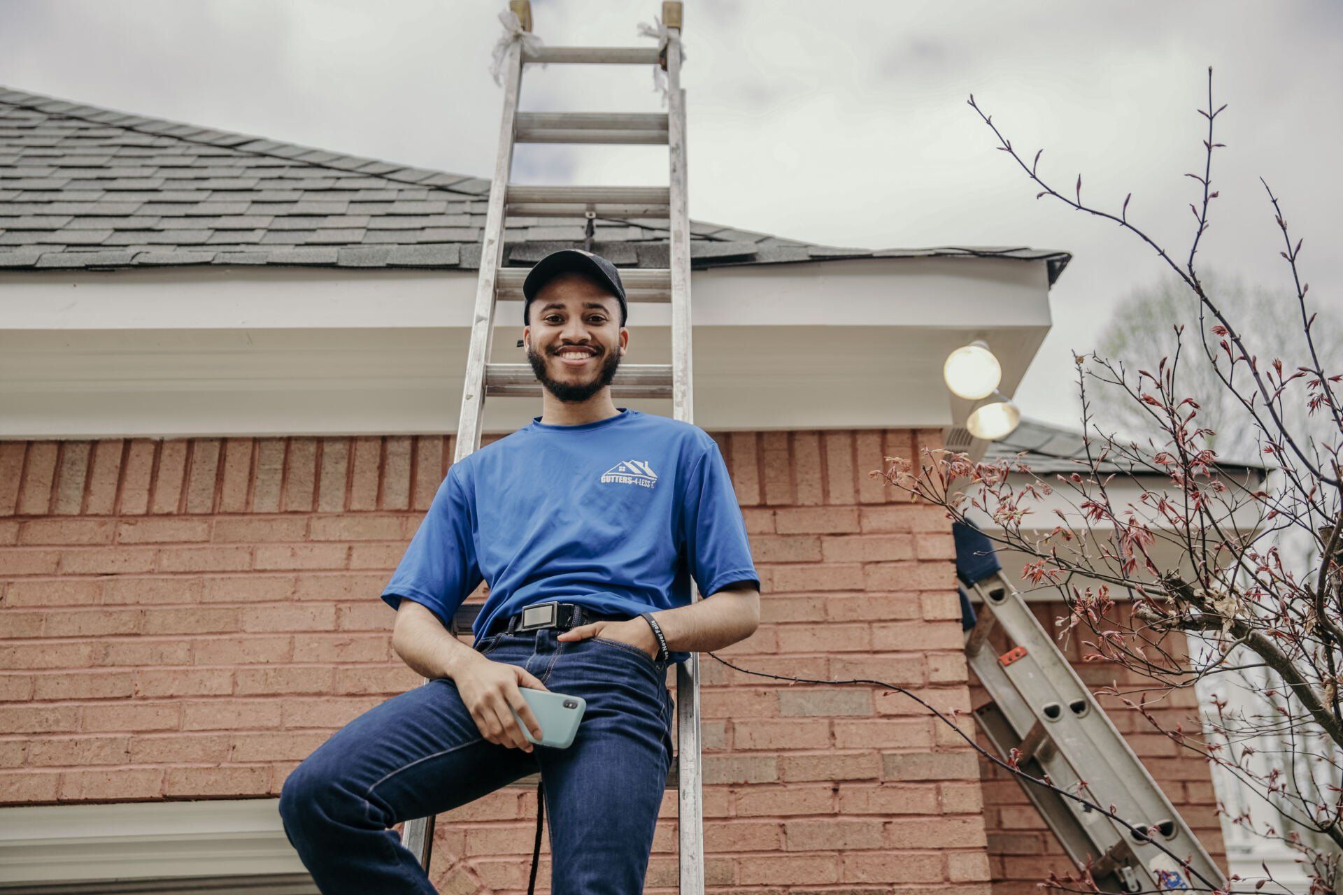 Person in blue shirt and jeans sitting on a ladder beside a brick house under repair