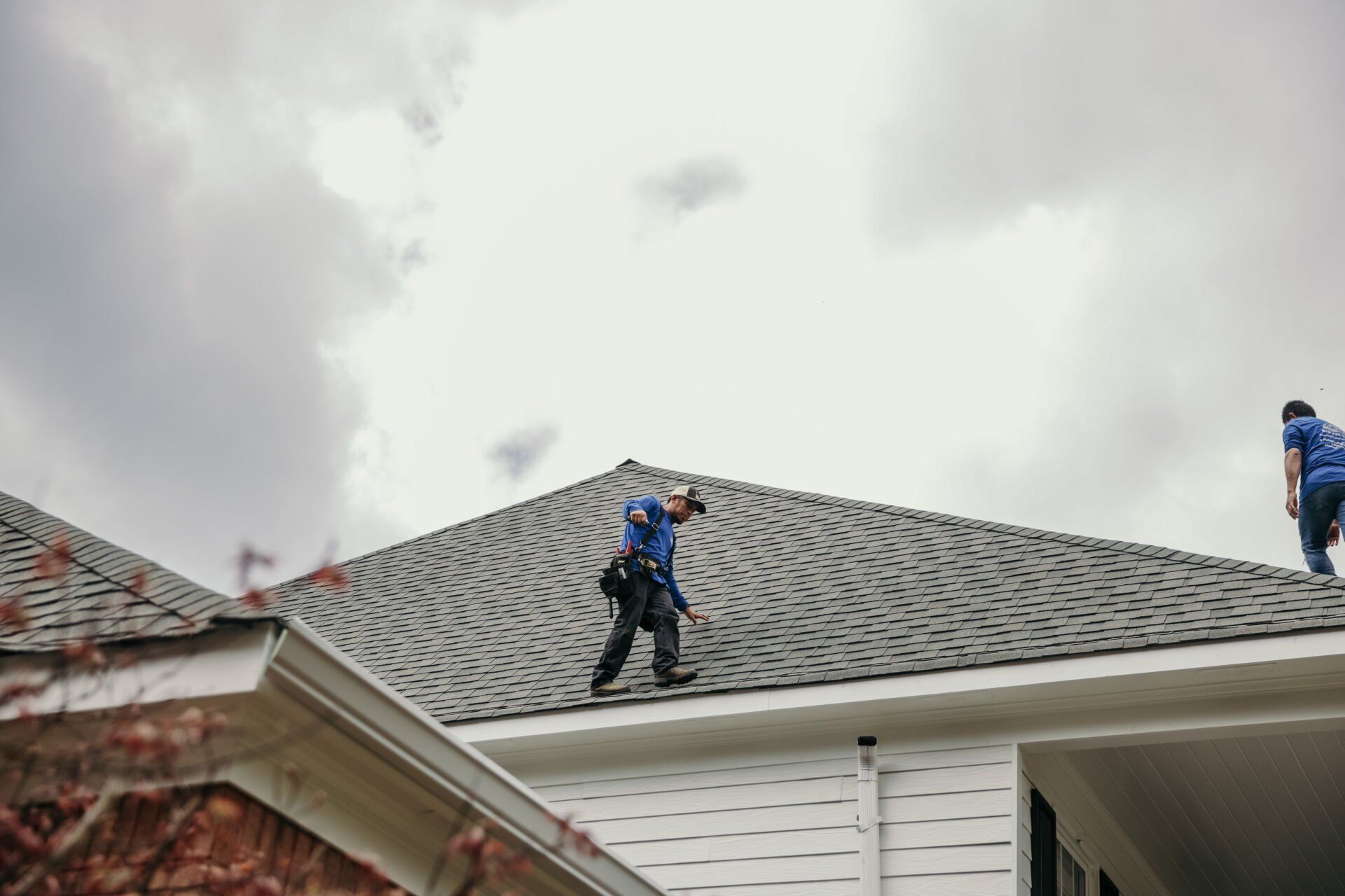 Two workers in blue uniforms walk across the gray shingled roof of a house under a cloudy sky.