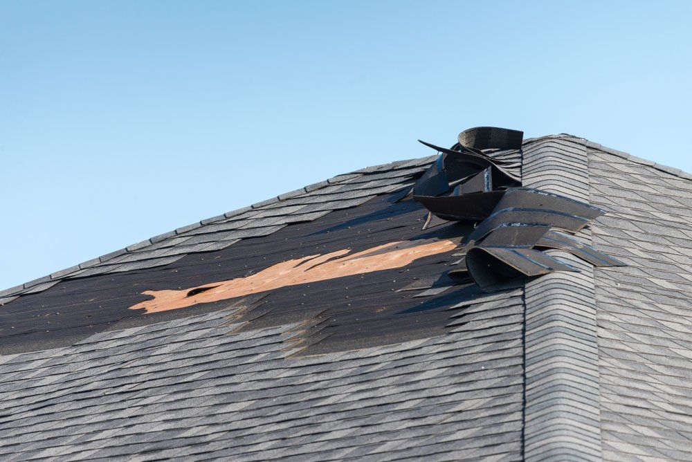 Roof shingles torn off near a vent, exposing damaged underlayment against a blue sky