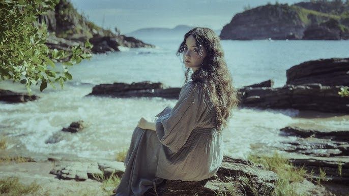 Woman seated on rocky shore, looking toward the sea, with cliffs and blue water in the background