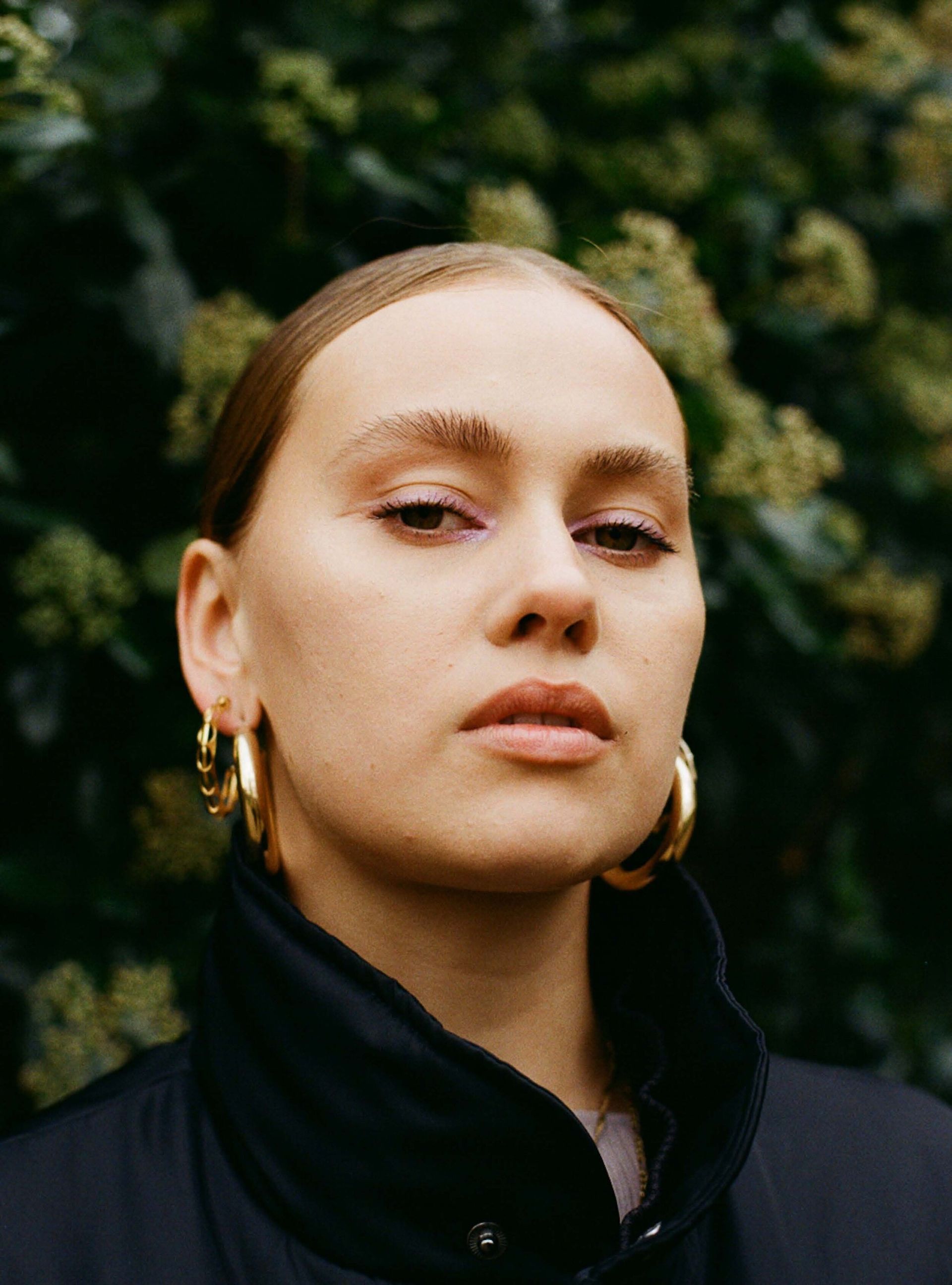 Portrait of a person in a dark jacket with gold hoop earrings, standing before leafy greenery