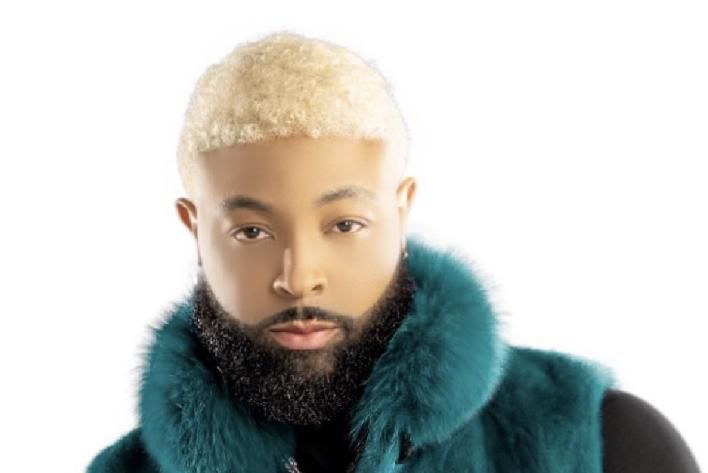 Portrait of a person with bleached hair, full beard, and teal fur-collared coat against a white background