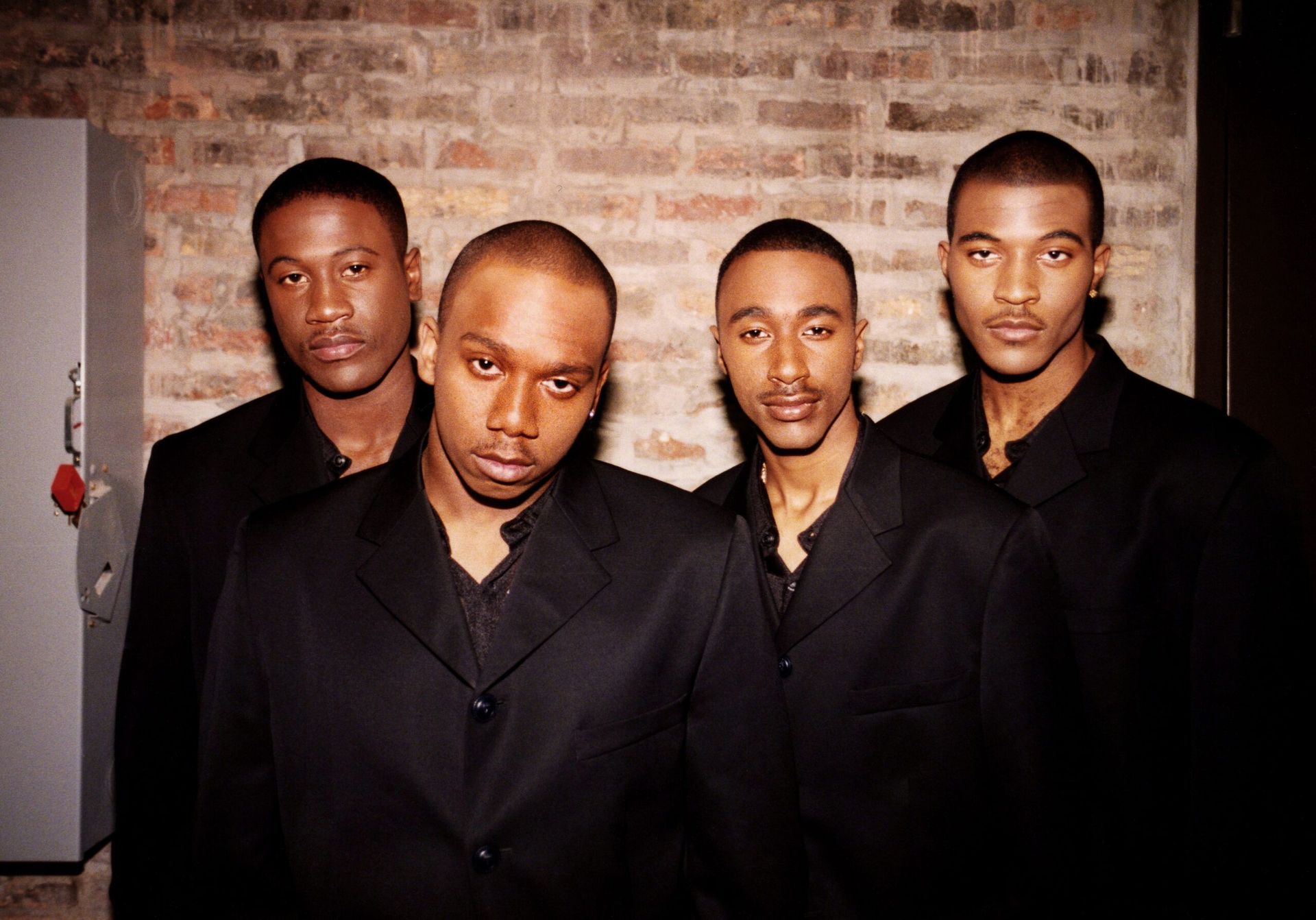Four men in black suits posing against a brick wall, looking directly at the camera.