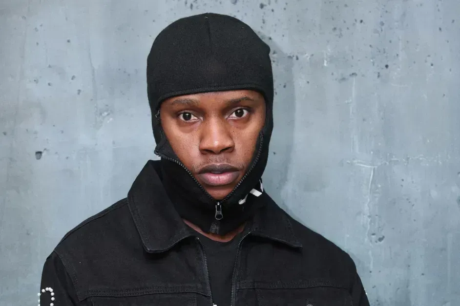 Person in black jacket and cap, looking at the camera against a gray concrete wall.