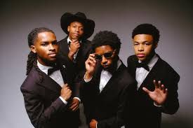 Four men in black suits pose together against a dark studio background, one adjusting his glasses.