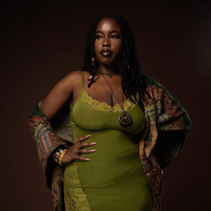 Woman in an olive-green dress and patterned shawl, standing in a studio pose against a dark background.