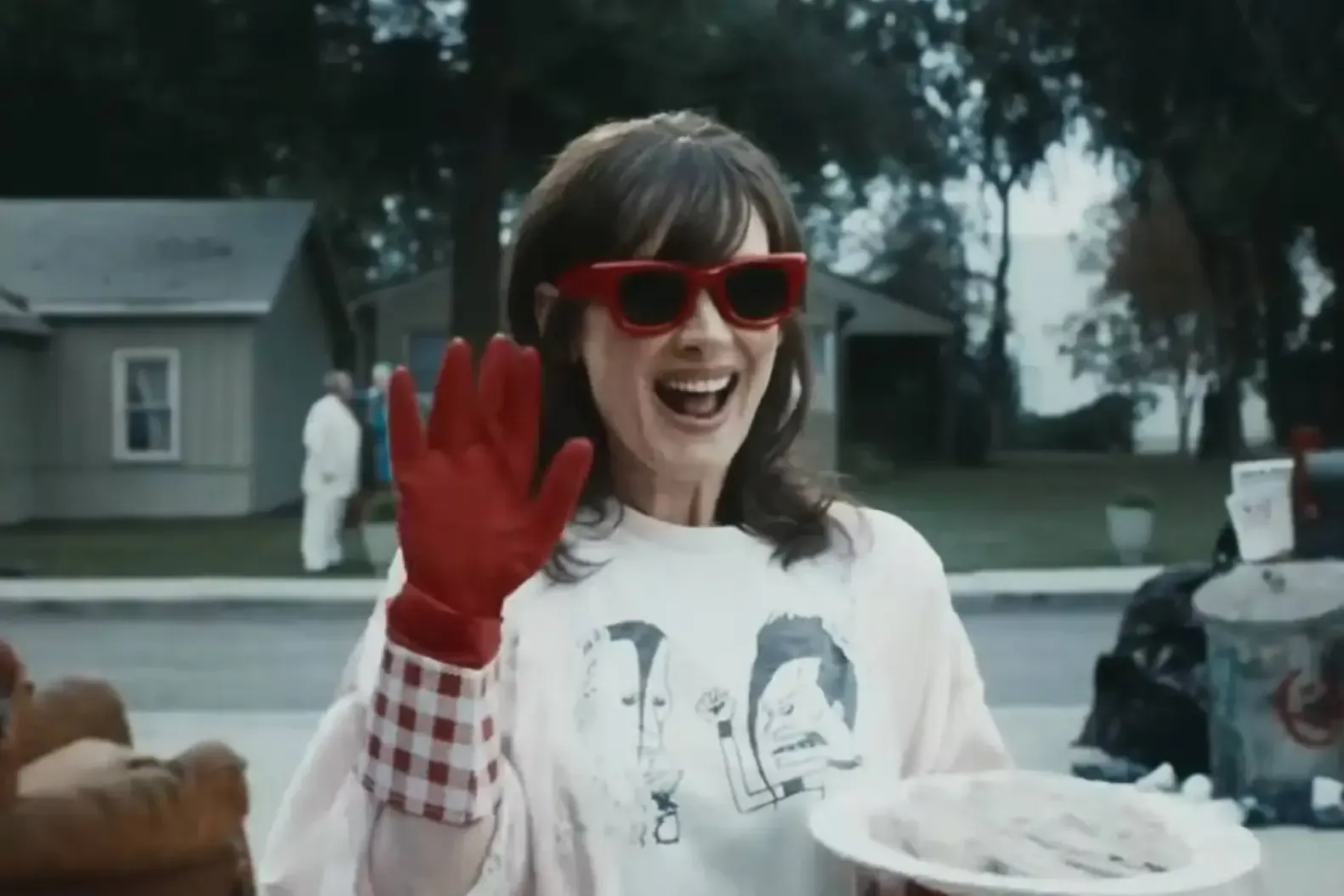 Smiling person in red sunglasses and gloves, waving while holding a plate outdoors in front of a house