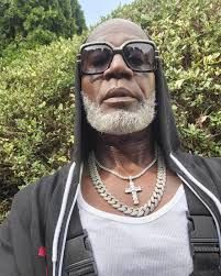 Man wearing sunglasses, silver chains, and black hoodie outdoors with green trees in the background