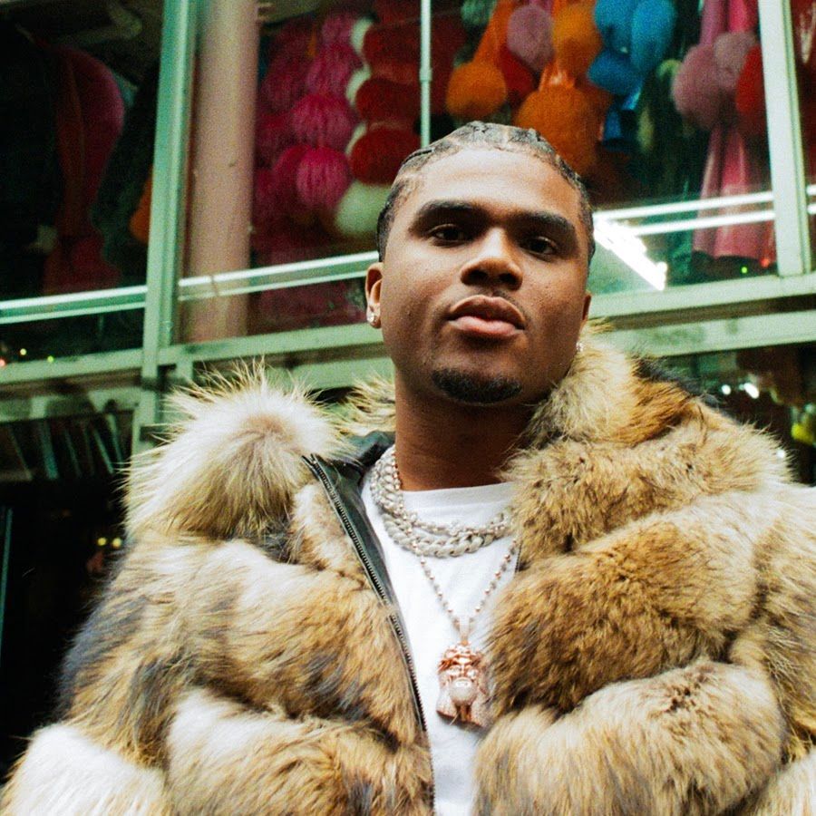 Person in a fur coat and chain necklace standing indoors with colorful hanging decorations in the background