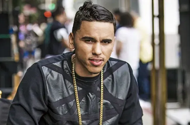 Man wearing a black jacket and gold chains standing on a city street, looking at the camera