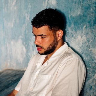 Man in a white shirt with a beard, seated in profile against a textured blue wall.