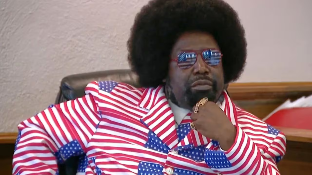 Man in red-and-white striped suit, blue tie, and mirrored sunglasses, reclining on a couch with hand on chin.