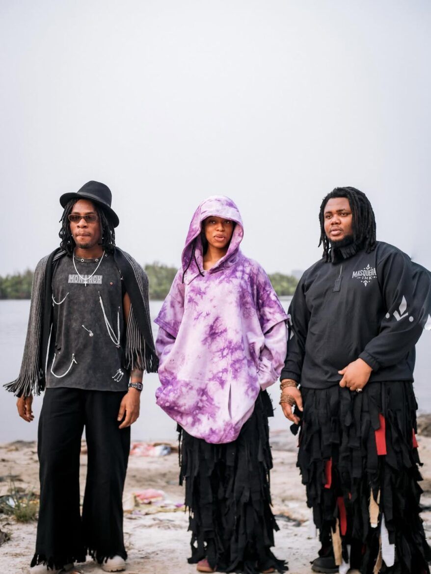 Three people in dark clothing and a purple hooded cloak stand on a rocky shoreline.