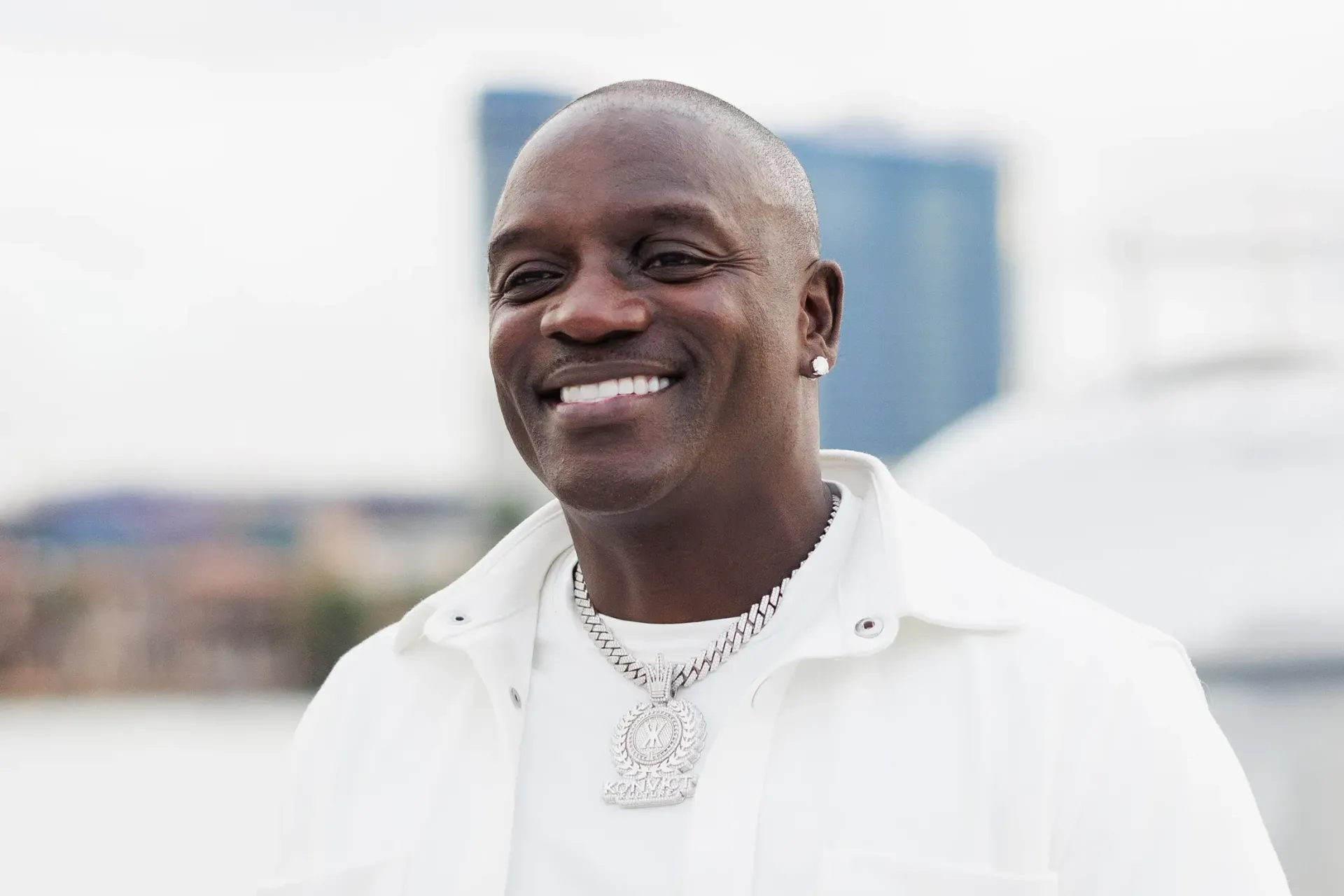 Smiling man in a white jacket and silver necklace outdoors with a blurred city skyline behind him