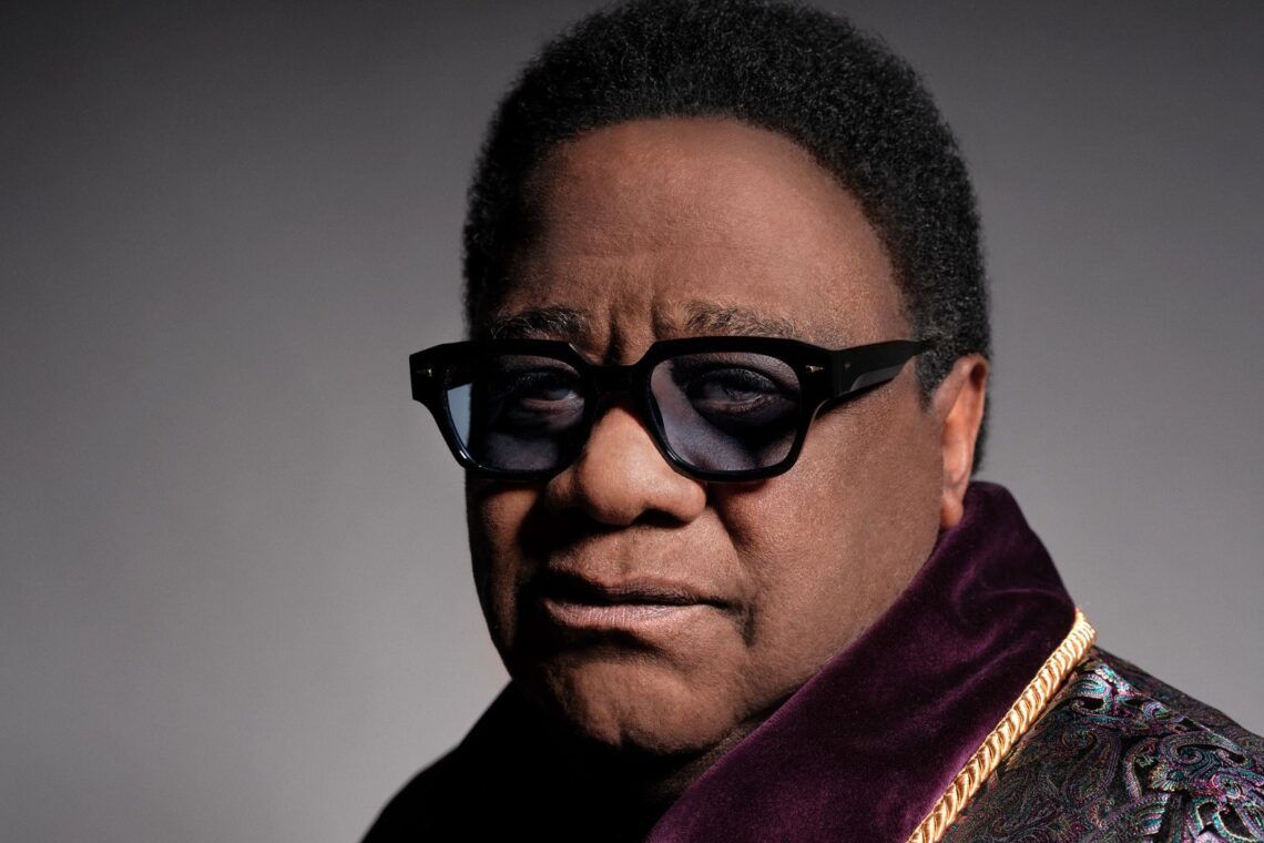Portrait of a man in black sunglasses and a dark patterned jacket against a gray background