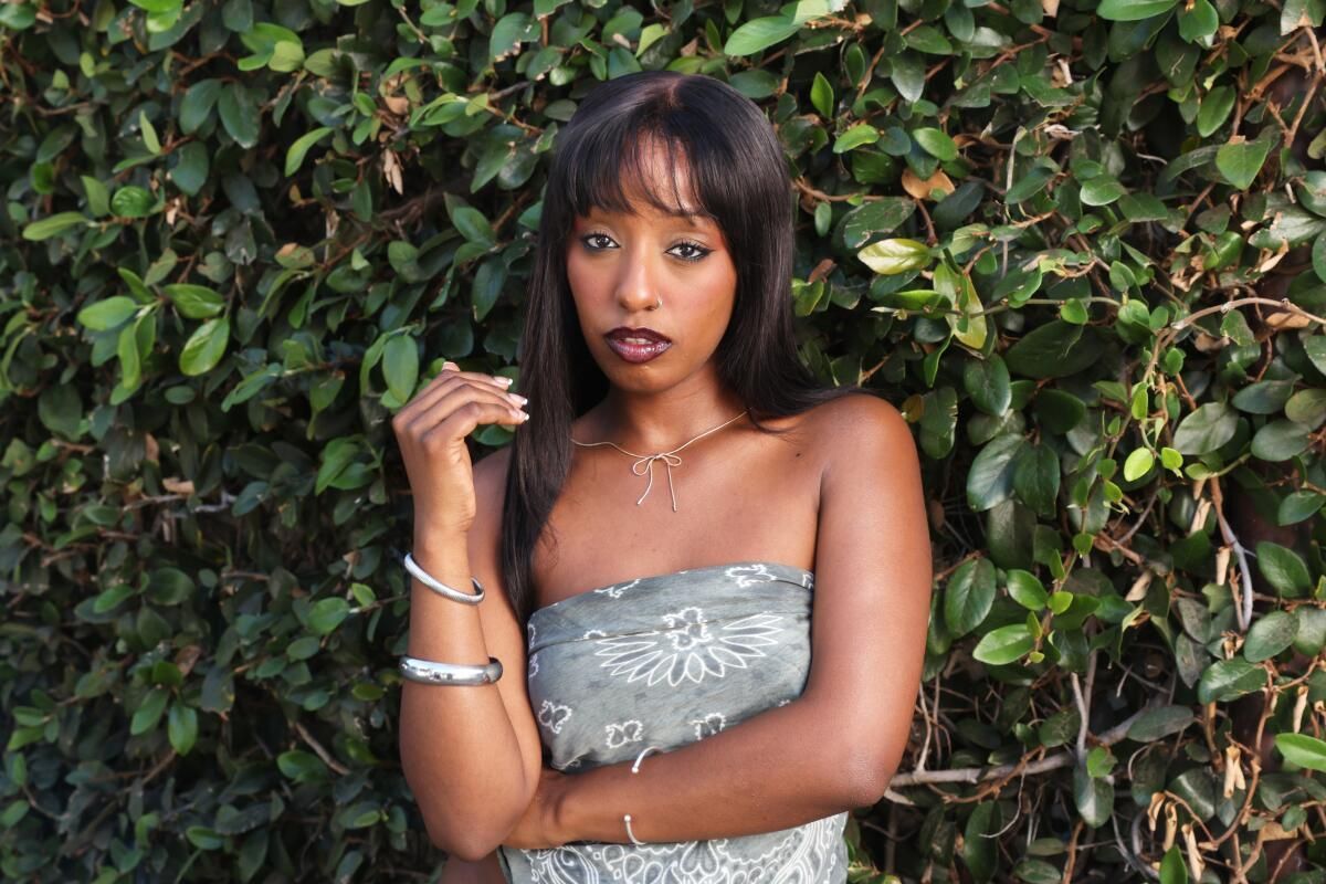 Woman in a silver top posing against a leafy green wall, looking at the camera.