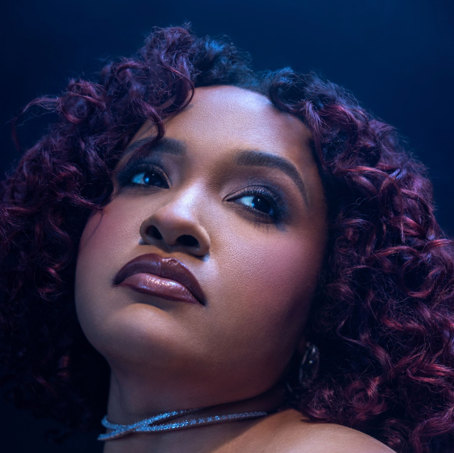 Close-up portrait of a person with curly hair and dark lipstick, gazing upward in blue-purple light