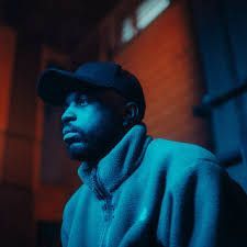 Blue-lit person in a cap and sweater standing against a red brick wall at night
