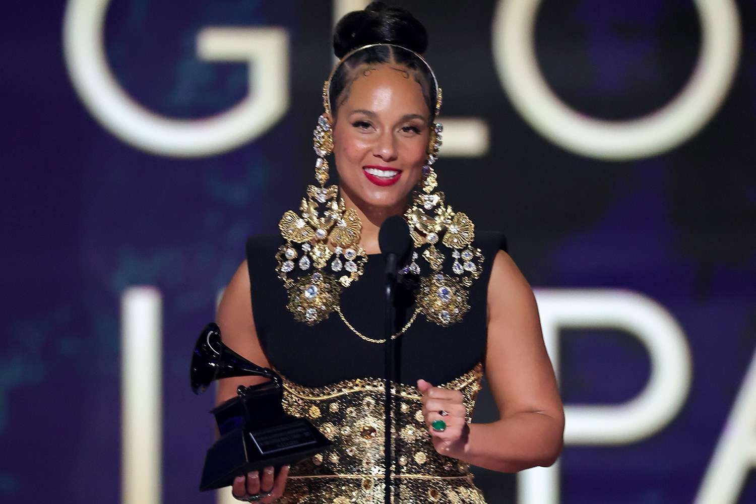 Woman in a black-and-gold gown holding a trophy on a red carpet, smiling at a gala event.