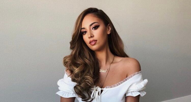 Woman with long wavy brown hair in an off-shoulder white top against a plain wall