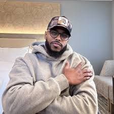 Man in a gray hoodie and cap sitting on a couch, arms crossed, in a softly lit living room.