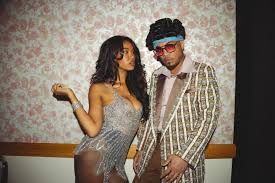 Two people posing indoors, one in a silver dress and the other in a striped suit with red sunglasses.