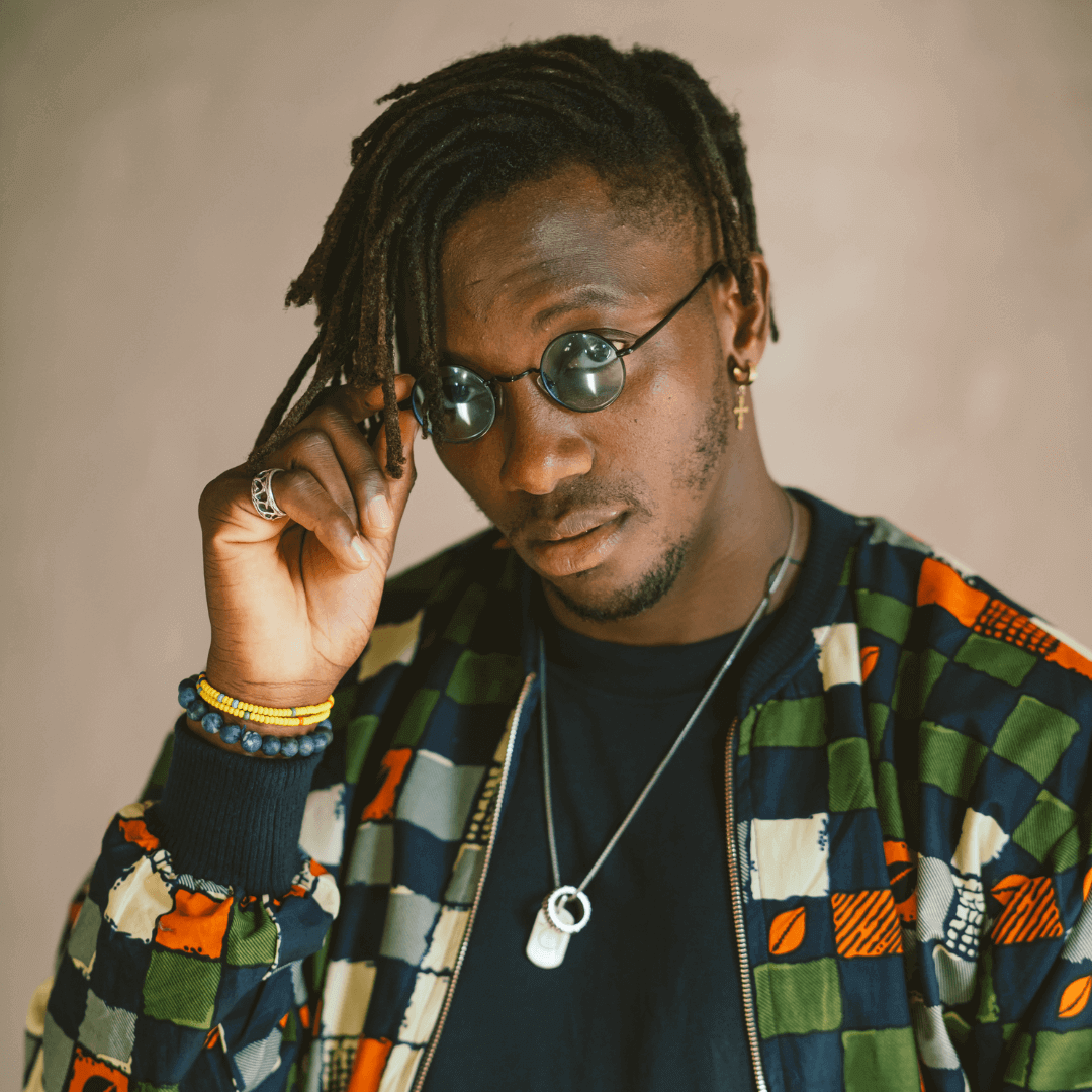 Man in sunglasses adjusting his hair, wearing a colorful patterned jacket and necklace against a beige background