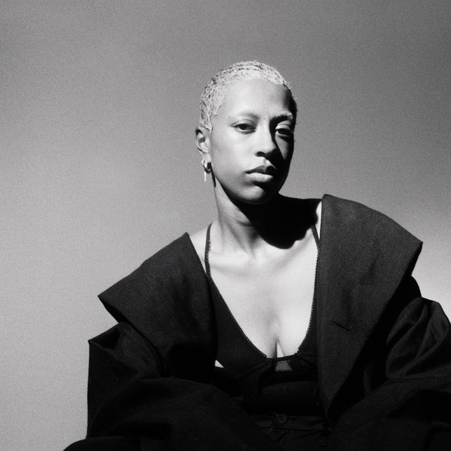 Black-and-white portrait of a person in a dark outfit with a neutral expression against a plain backdrop