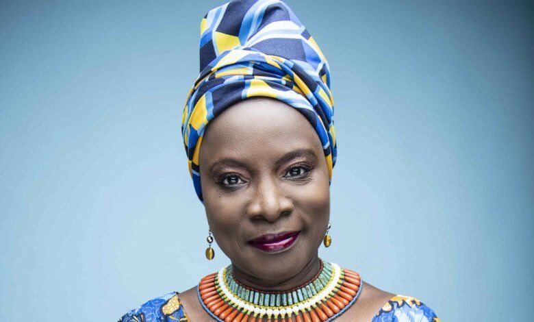 Portrait of a woman wearing a blue-and-yellow headwrap and colorful necklace, smiling against a light blue background