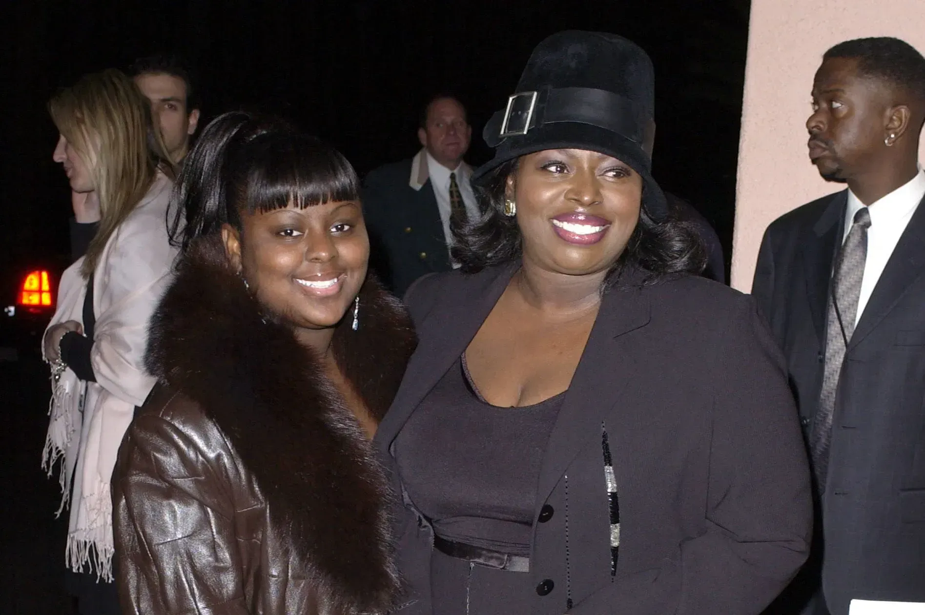 Two women smiling at a nighttime event, one in a fur-trimmed coat and the other in a black hat and dark outfit