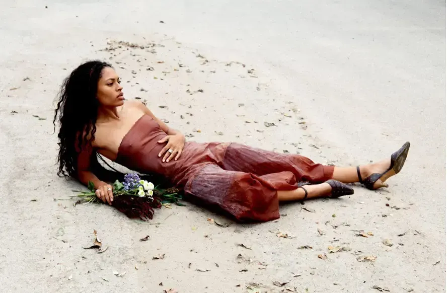 Woman reclining on a sandy beach beside flowers, wearing a maroon dress and black heels