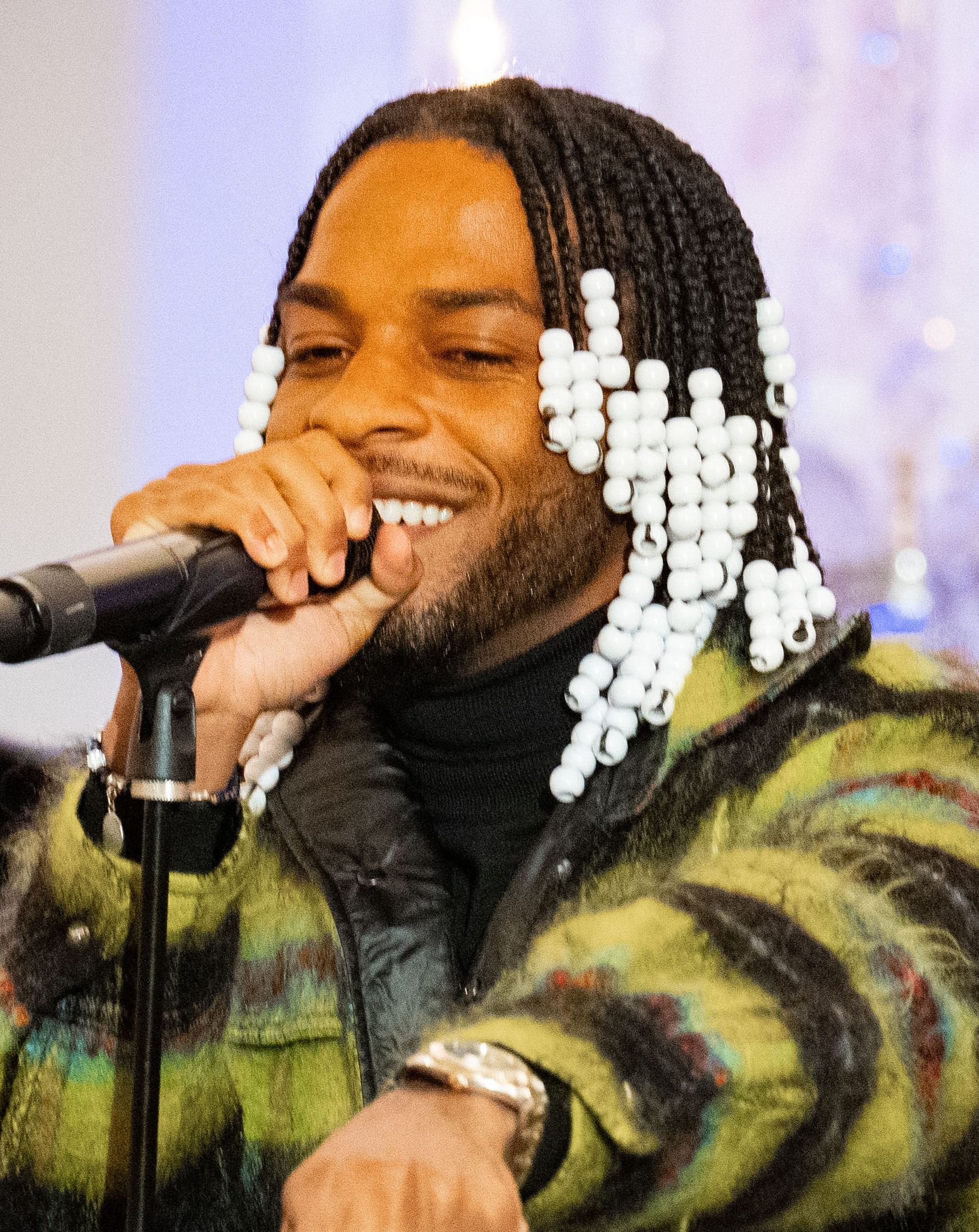Singer smiling into a microphone, wearing a patterned jacket and beaded braids, mid-performance