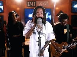 Three musicians performing in a studio, with a singer at a microphone and guitarist behind him.