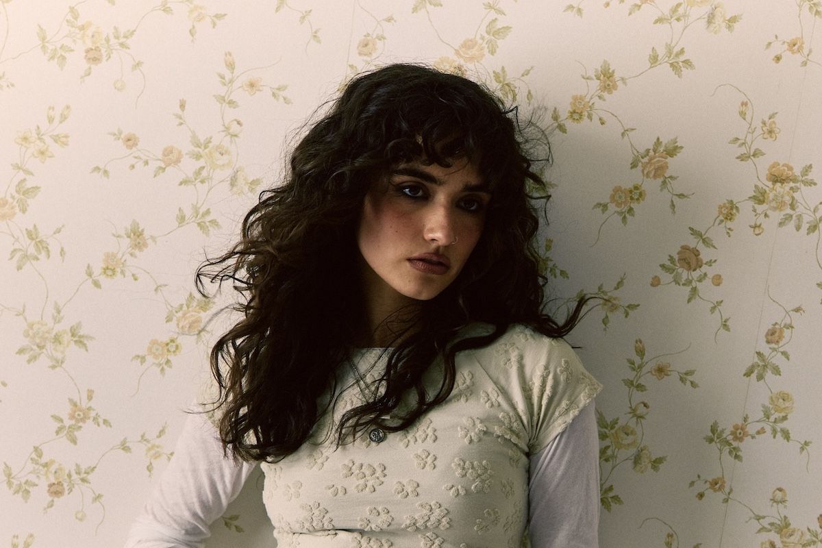 Woman with dark curly hair in a cream top, posing against floral wallpaper.