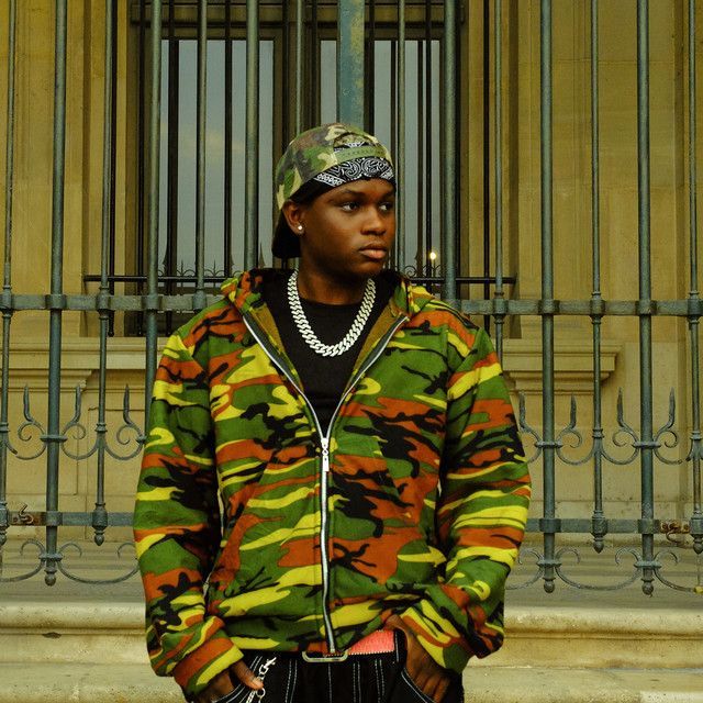 Person in a camouflage jacket and bandana standing before ornate iron bars and a stone wall.