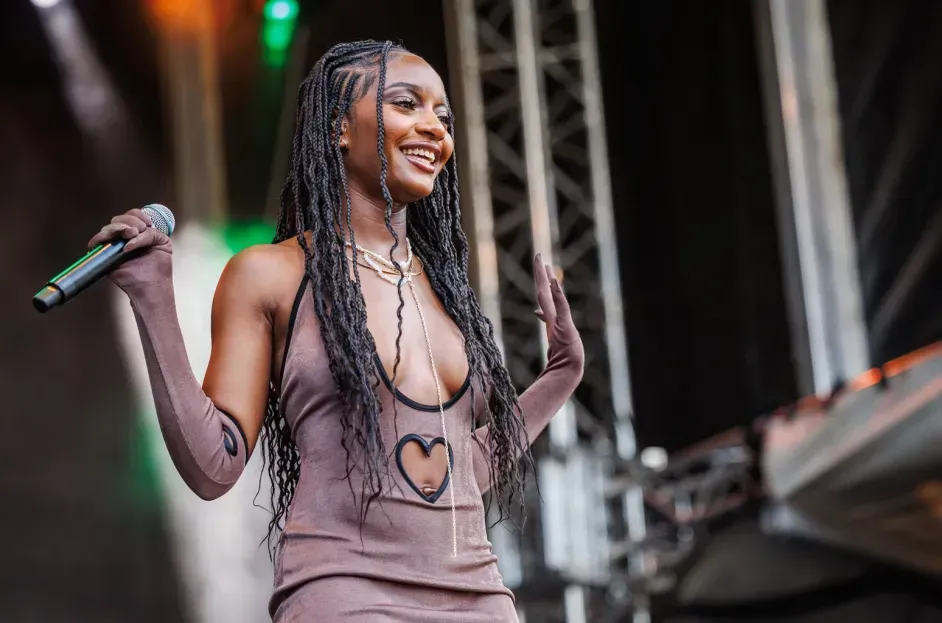 Singer holding a microphone on stage, smiling and wearing a tan dress with long braided hair