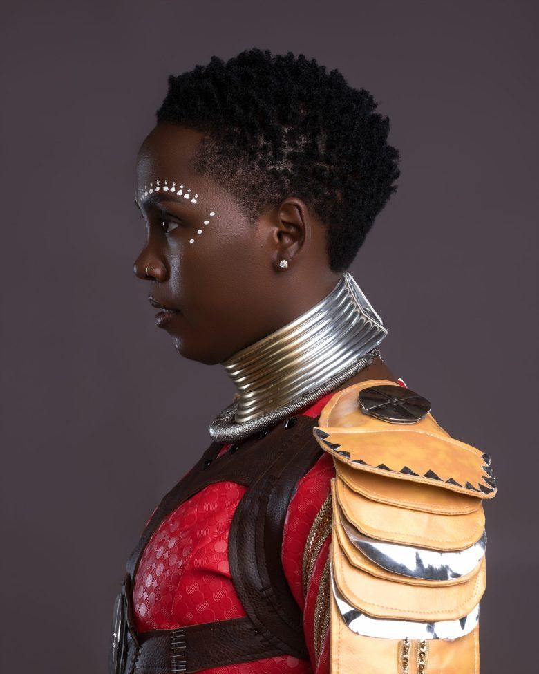 Profile portrait of a person in a red outfit with metallic neck rings and orange shoulder accents