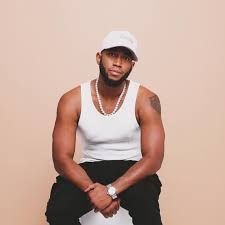 Man in white tank top and cap seated against a beige background, wearing a silver chain and watch.