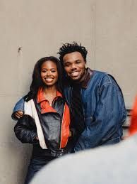 Two smiling people posing together against a plain wall, one in a black-and-orange jacket and one in a blue jacket.