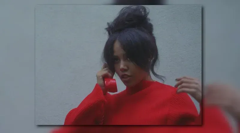 Woman in a bright red sweater adjusting her hair in front of a plain gray wall